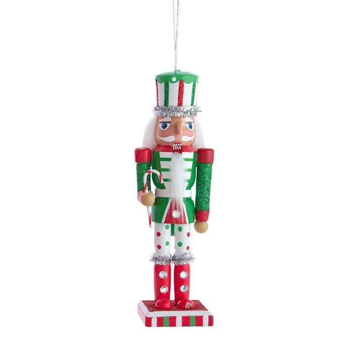 Red, green, and white nutcracker ornament with glittered accents, white beard, polka dot leggings, and a small candy cane on a striped rectangular base.