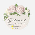 Round white ornament with pink and white roses, green leaves, and text 'Bridesmaid I can't say I do without you' on a white background. Can be personalized