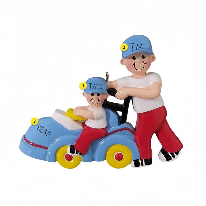 Decorative ornament of a man pushing a child on a toy car with example names and year text.