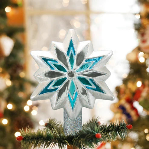 Silver, white, and blue snowflake tree topper displayed on top of a decorated Christmas tree branch with lights in the background.