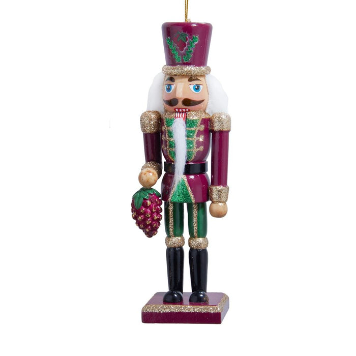 Burgundy, green, and gold nutcracker ornament with a white beard, glittered trim, and a purple grape cluster in hand on a rectangular base.